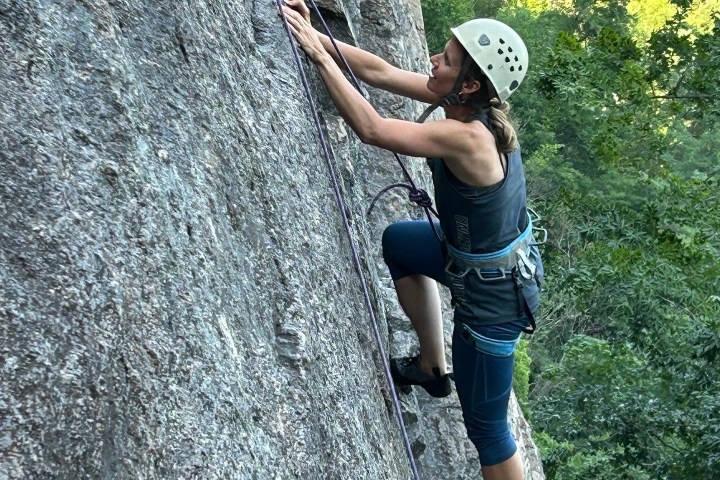 Person rock climbing on a steep cliff wearing a helmet and harness, surrounded by greenery.