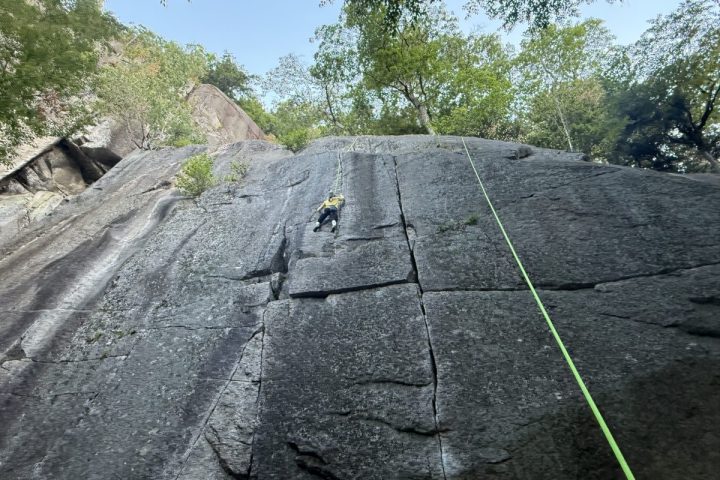 Rock climber ascending a steep cliff with a green rope under a clear sky and surrounded by trees.