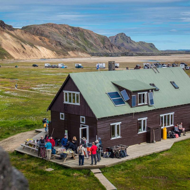 People gathered near a wooden house with mountains in the background and parked cars in an open field.